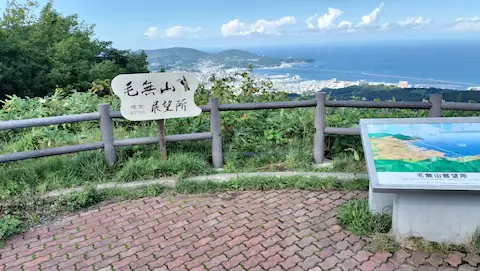 毛無山展望台の全景。眺望を説明した看板と、毛無山展望所と書かれた木の看板がある展望台から、小樽の街並みと海が見える。